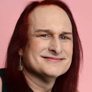 A person with long hair and earring smiles against a pink background, looking directly at the camera. No recognizable landmarks or buildings visible.