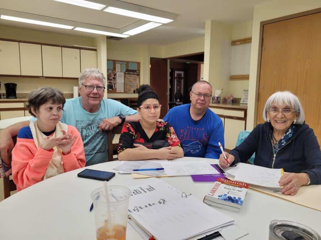 Five people sitting at a table with papers and a Spanish book, likely engaging in a study session in a room with cabinets.