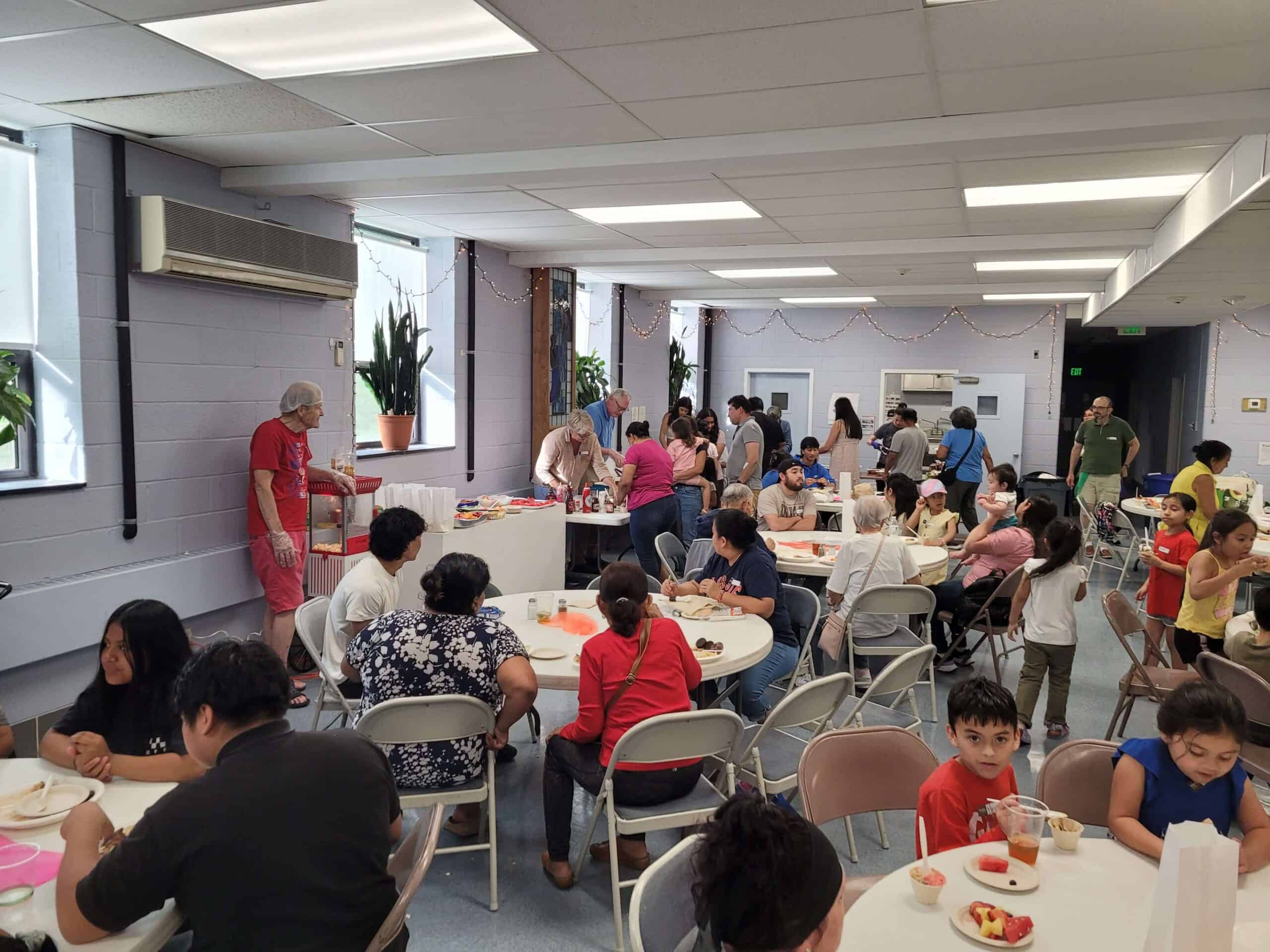 A community gathering in a bright room with people eating and socializing at tables, featuring decorations and refreshments in a relaxed atmosphere.
