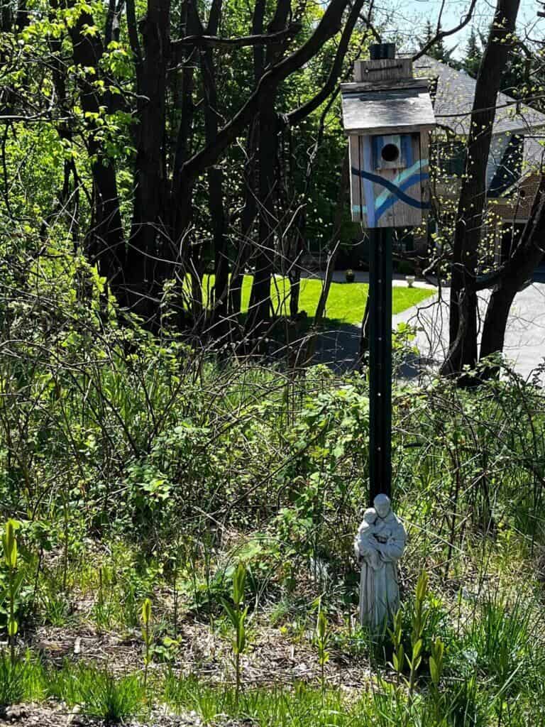 A painted birdhouse on a post amidst trees with a small religious statue below. Sunlit greenery surrounds the area, creating a serene atmosphere.