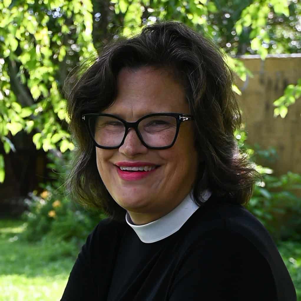 St. Francis Episcopal Parish & Community Center A smiling person with glasses and a clerical collar is outdoors, surrounded by lush greenery and sunlight filtering through the trees.