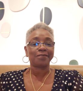 A person with glasses and hoop earrings smiles at the camera against a decorative wall with abstract shapes in a cozy setting.