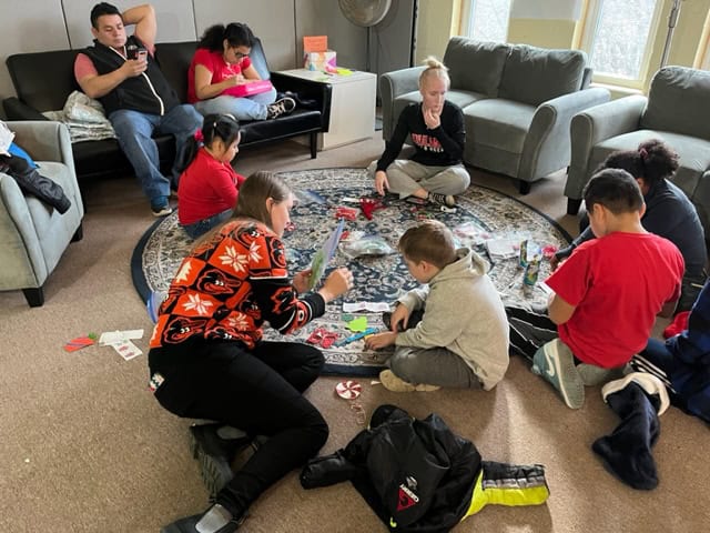 People and children gather in a cozy living room, engaged in arts and crafts, sitting on a decorative carpet among comfortable sofas.