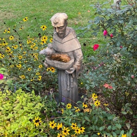 A garden statue of a hooded person holding a bowl, surrounded by vibrant yellow and pink flowers, in a grassy area.