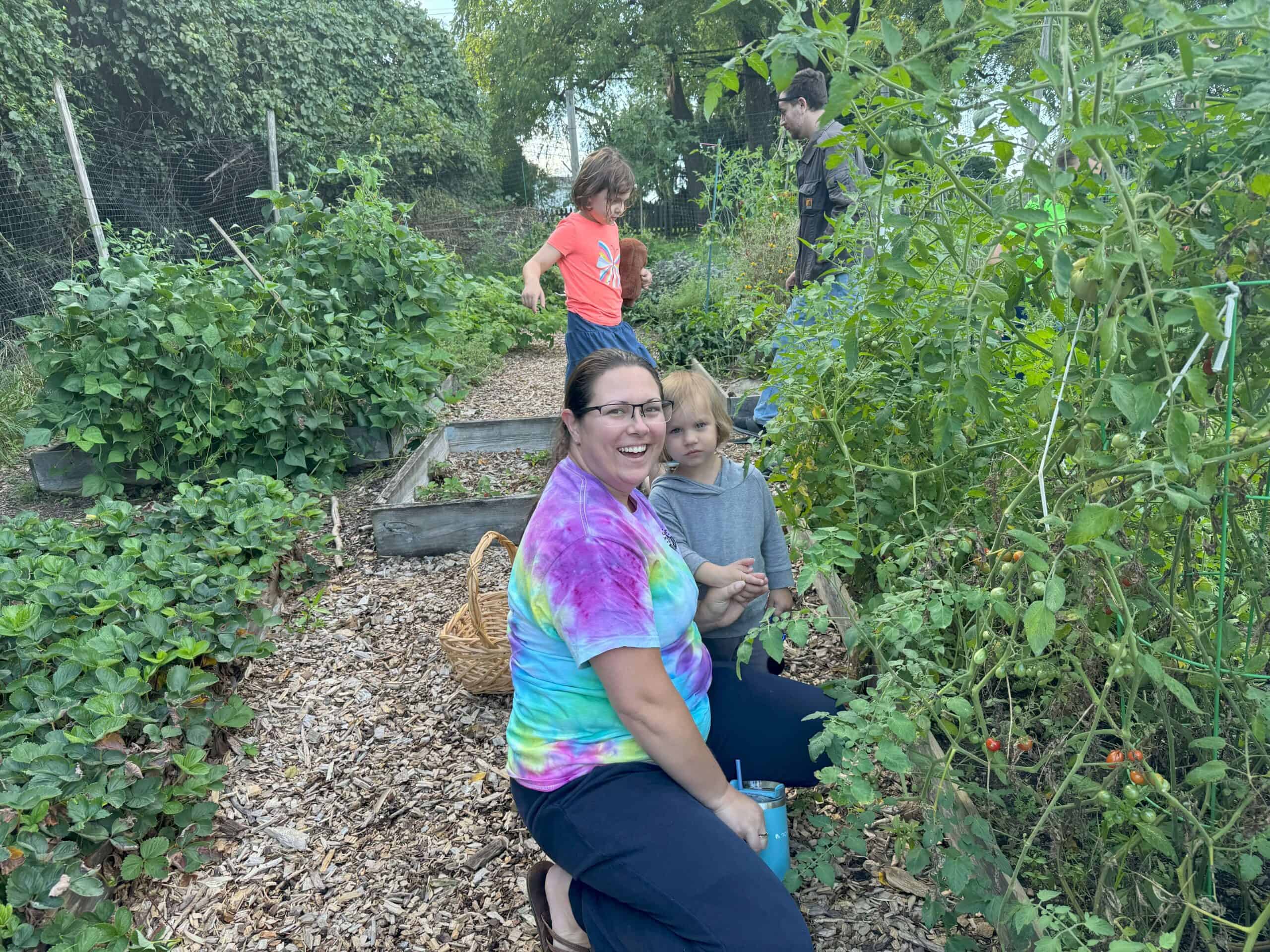 A group enjoying a thriving vegetable garden, one person holds a young child. Green plants surround them on a sunny day.