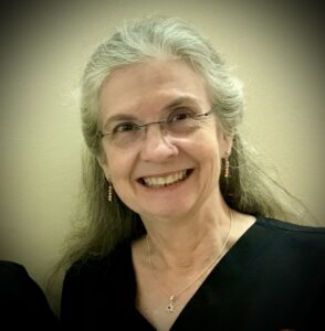 A smiling person with long gray hair and glasses, wearing a black top and necklace, stands against a plain indoor background.