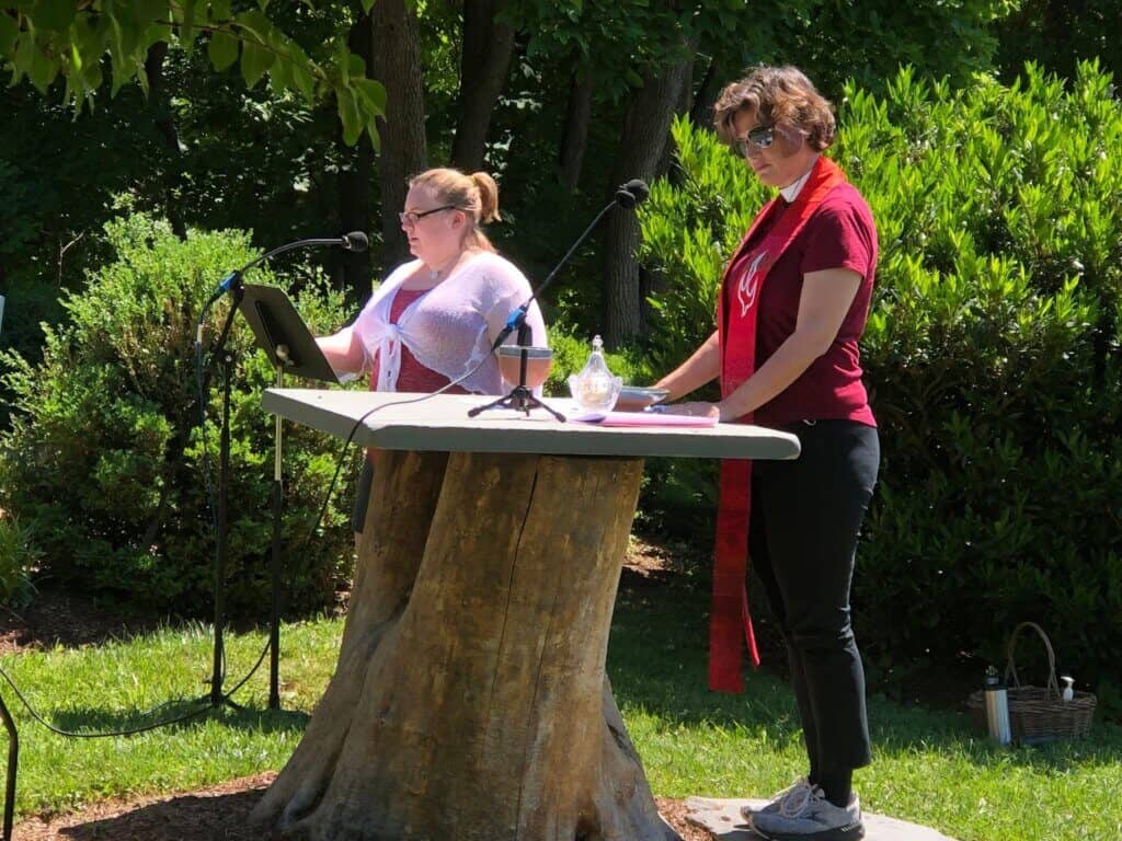Two persons stand at a podium outdoors, surrounded by greenery, reading or presenting with microphones. A basket is on the ground nearby.
