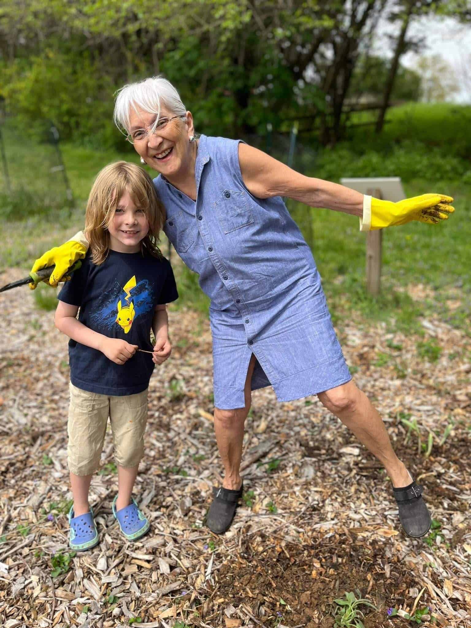 A person wearing yellow gloves and a child smiling outdoors, standing on wood chips with trees in the background, enjoying a garden setting.
