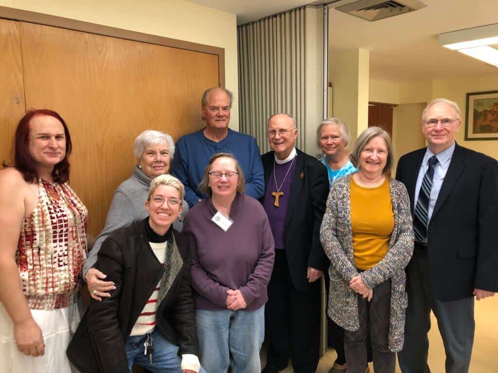 Nine people smiling indoors, standing together near a wooden door and partition. Casual and semi-formal attire; warm, friendly atmosphere.