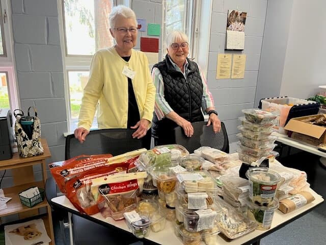 Two people stand behind a table full of assorted packaged food items in a bright room with gray brick walls and windows.