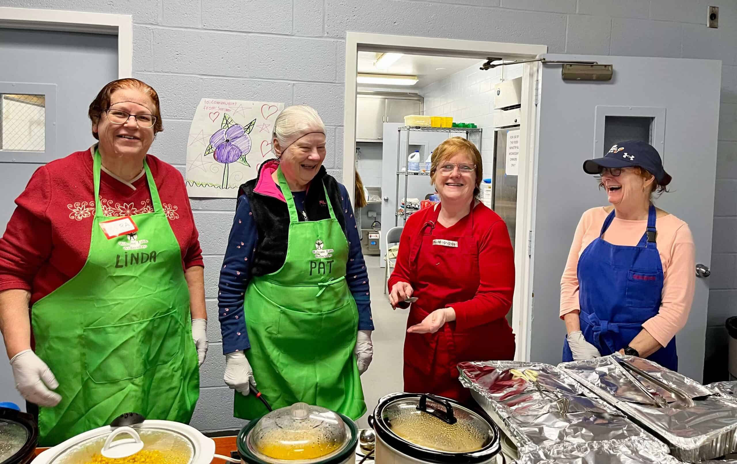Four people in aprons stand smiling in a kitchen, serving food from crockpots, with colorful drawings on the wall behind them.