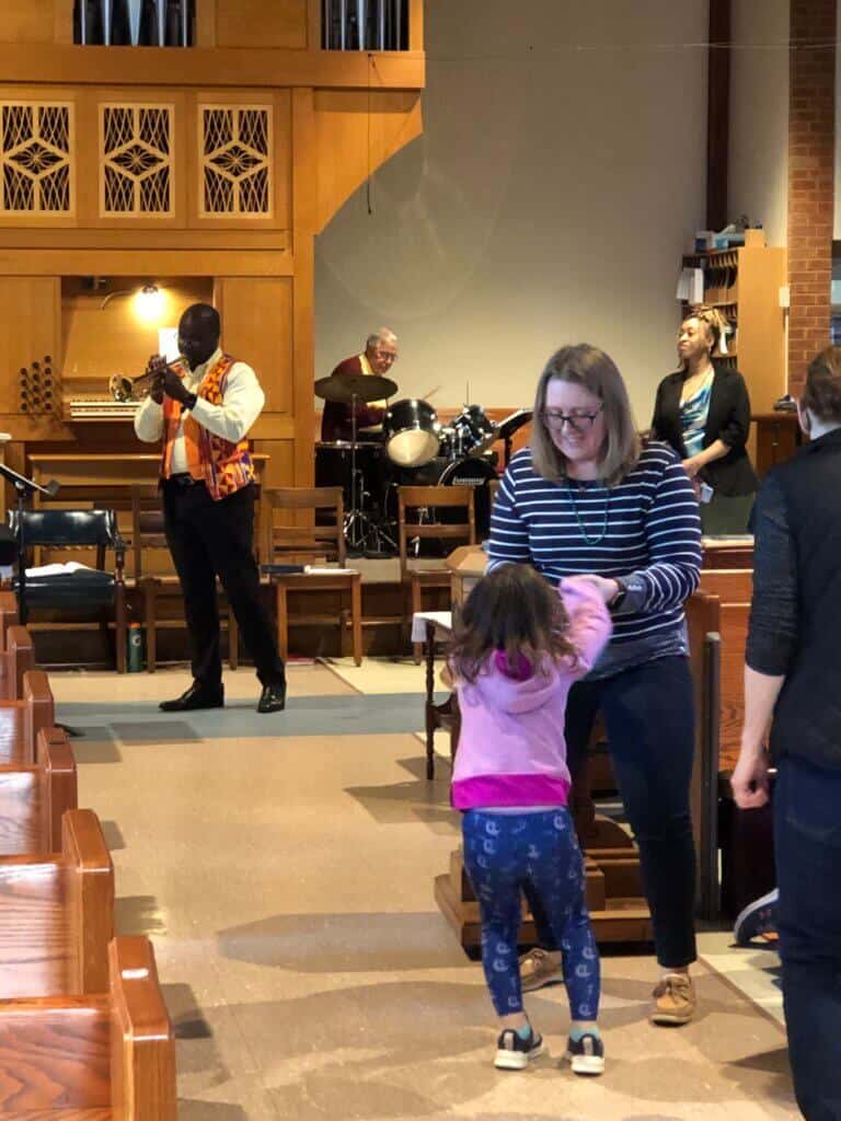 A person plays trumpet while another drums in a church setting. A person and child joyfully interact in the aisle.