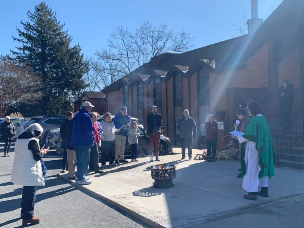 A group of people gathers around a fire outside a brick building, possibly a church, on a clear day.