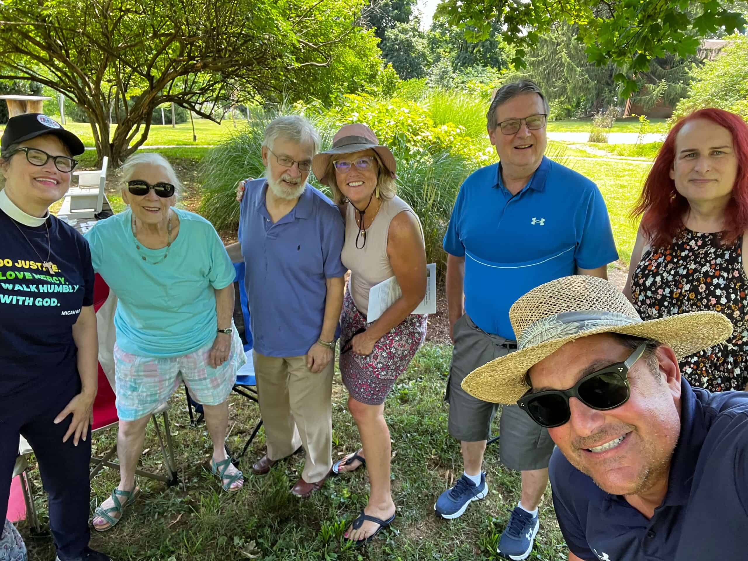 A group of seven people smiling outdoors in a lush, green park setting, with trees and grass in the background.