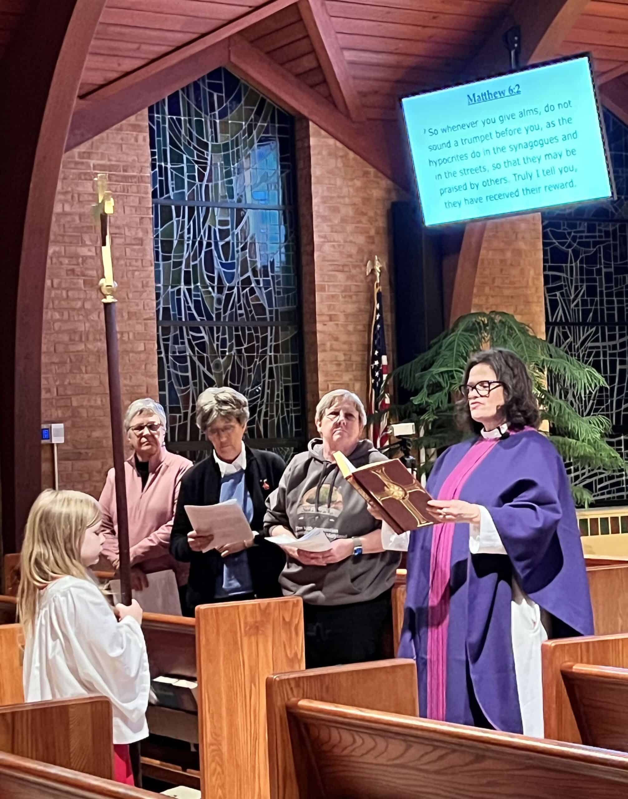 A child and four adults stand in a church interior, beneath a stained glass window. A screen displays a biblical passage.