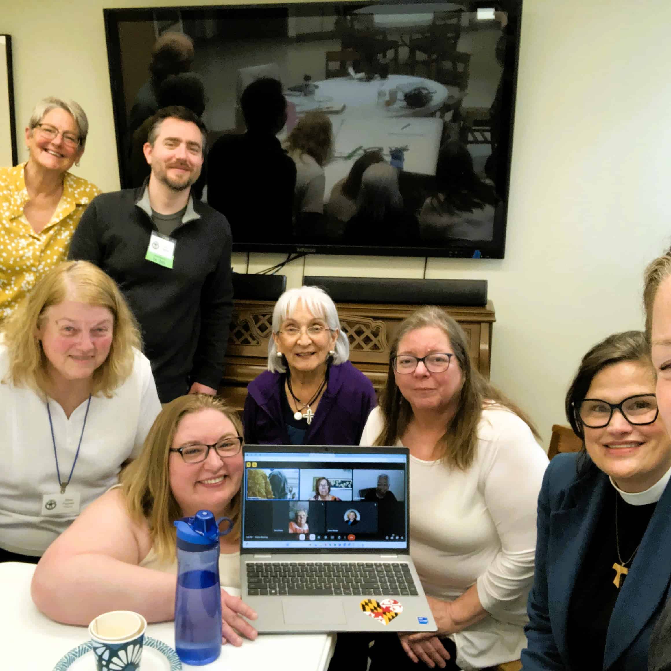 A group of seven people smiling, with one holding a laptop showing a virtual meeting, gathered in a room with a screen.