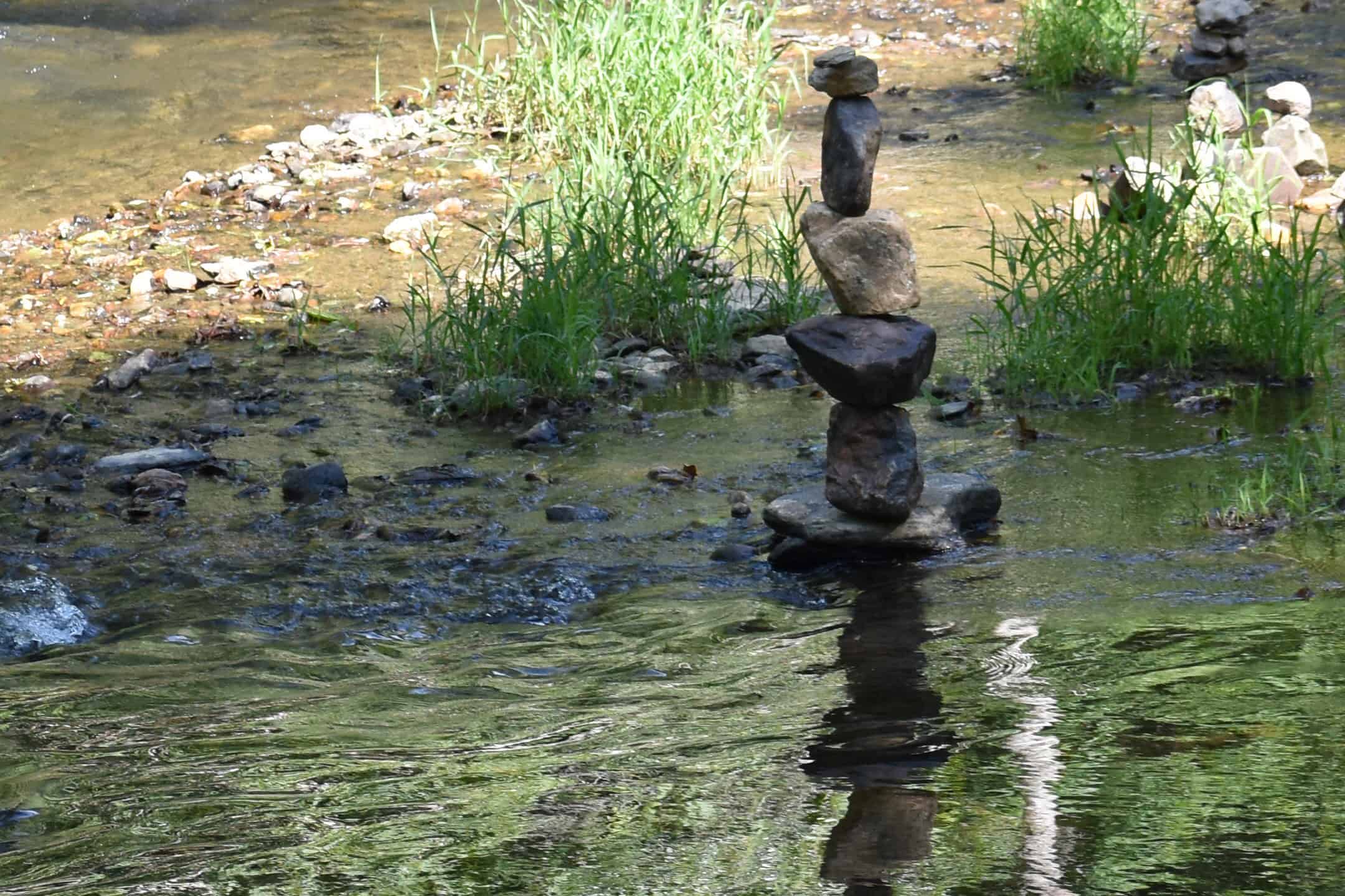 Balanced stones form a stack in a shallow, reflective stream, surrounded by small rocks and green plants. Peaceful natural scene with sunlight.