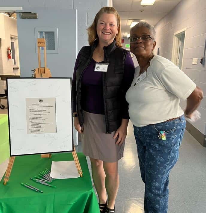 Two people smiling beside a signed document on an easel. Green pens and a tablecloth accent the setup in a hallway.