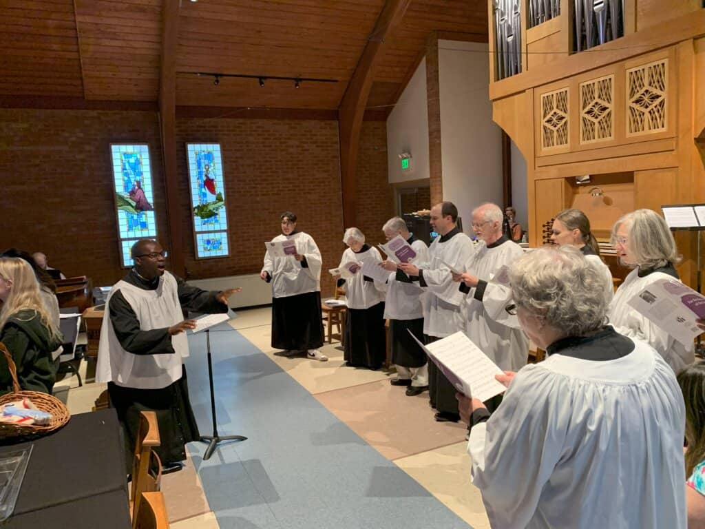 A choir sings in a church with stained glass windows and organ, led by a person in a white robe conducting.