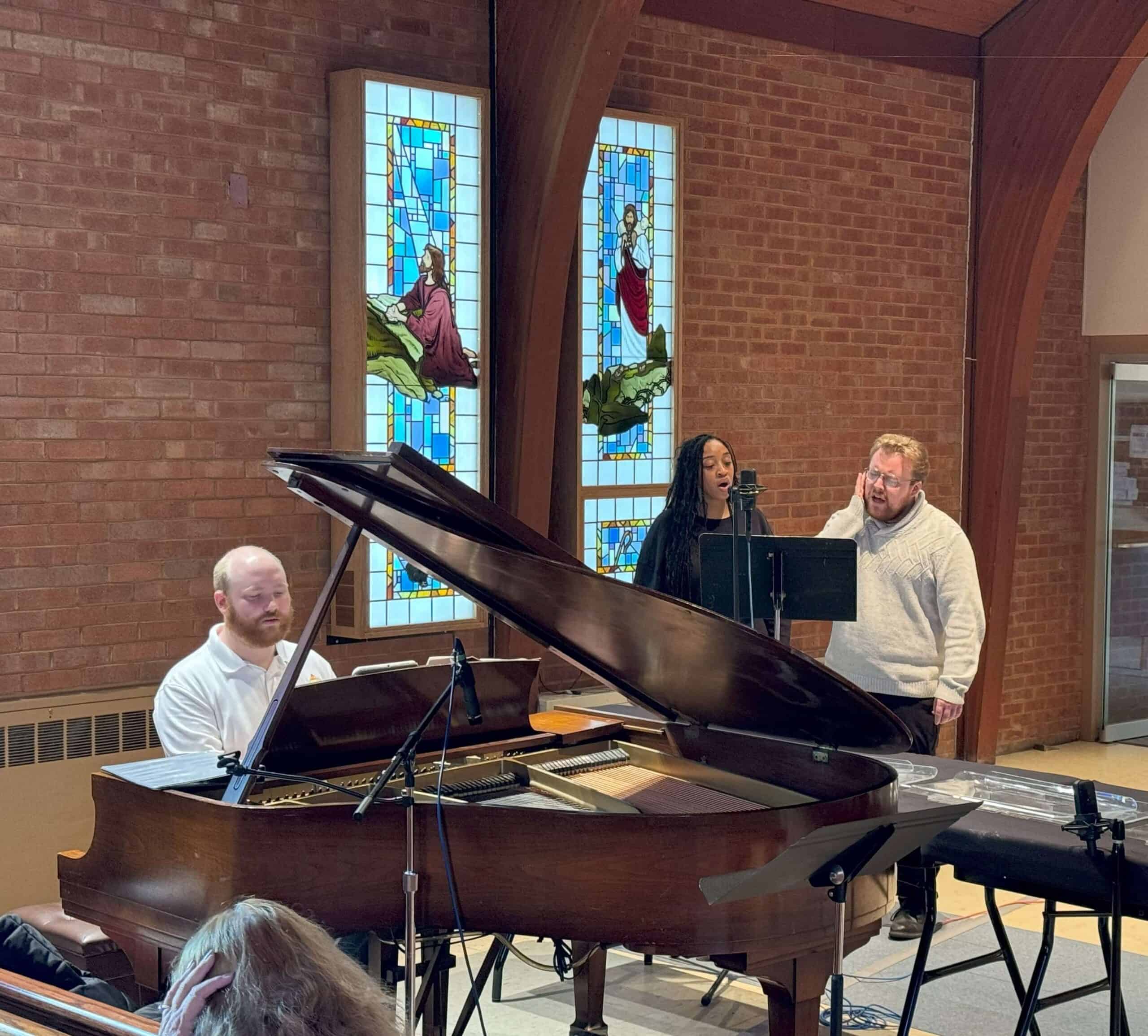 Three people perform in a room with stained glass windows. A person plays the grand piano while two sing beside it.