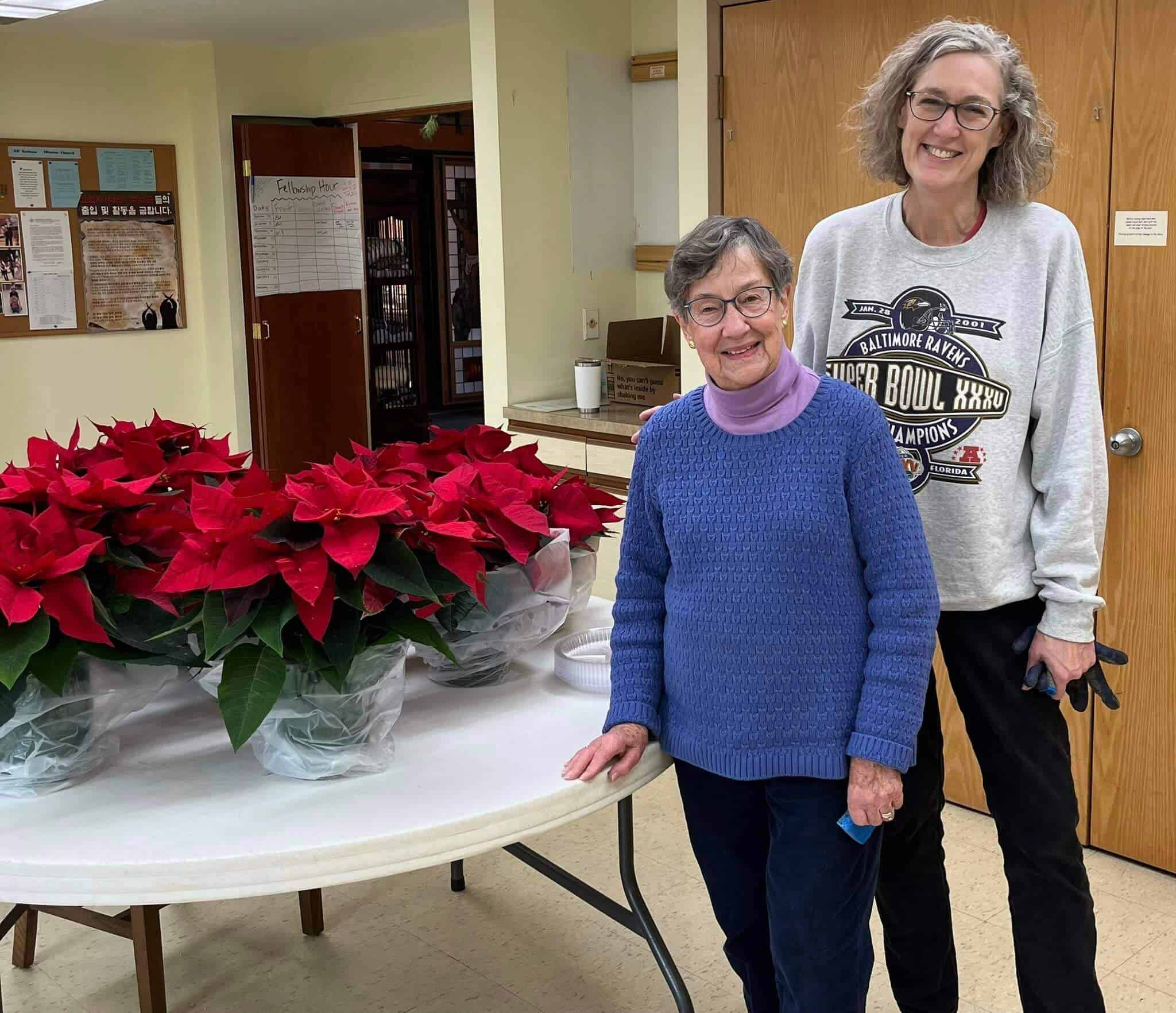 Two people smiling next to a table filled with poinsettias inside a room. A bulletin board is visible in the background.
