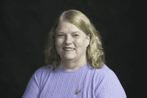 A person with long blonde hair wears a purple sweater, smiling against a dark background. There are no landmarks or buildings visible.