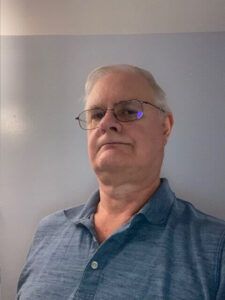A person wearing glasses and a blue shirt stands against a plain, light-colored wall, looking directly at the camera.