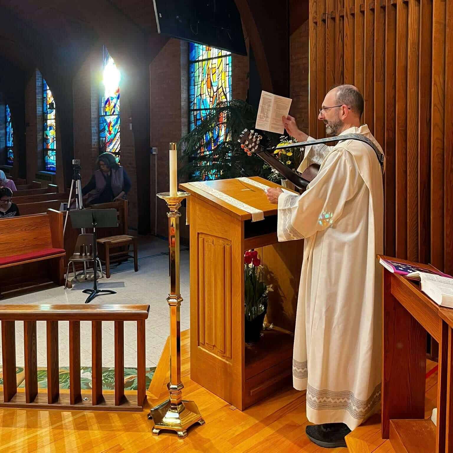A person dressed in religious robes plays guitar and sings in a church featuring stained glass windows and wooden architecture.
