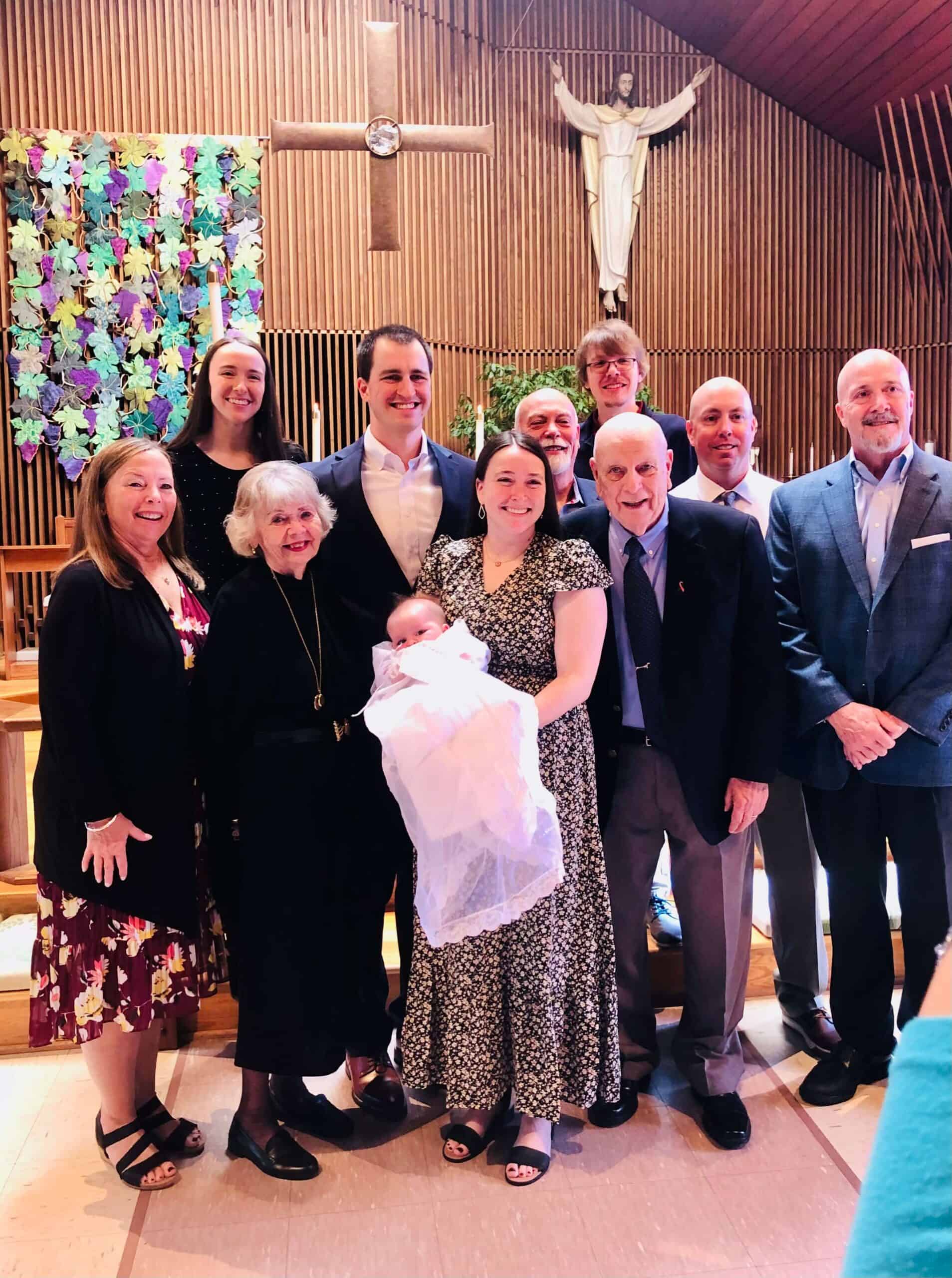 A group of people smiling with a baby, posing in a church setting featuring a wooden cross and colorful leaf artwork.