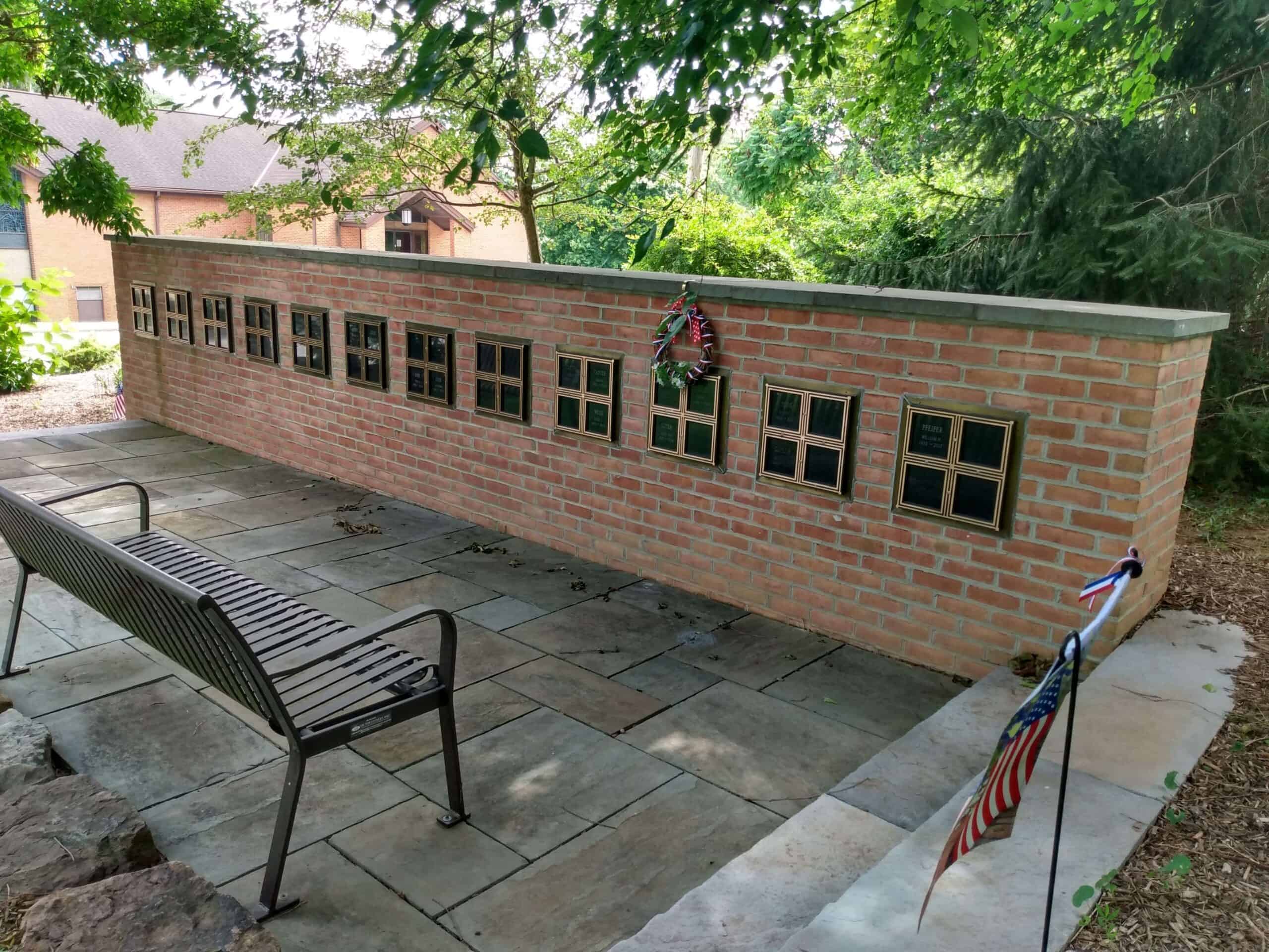A tranquil memorial area with a brick wall displays plaques, a bench, and flags, surrounded by trees and a nearby building.