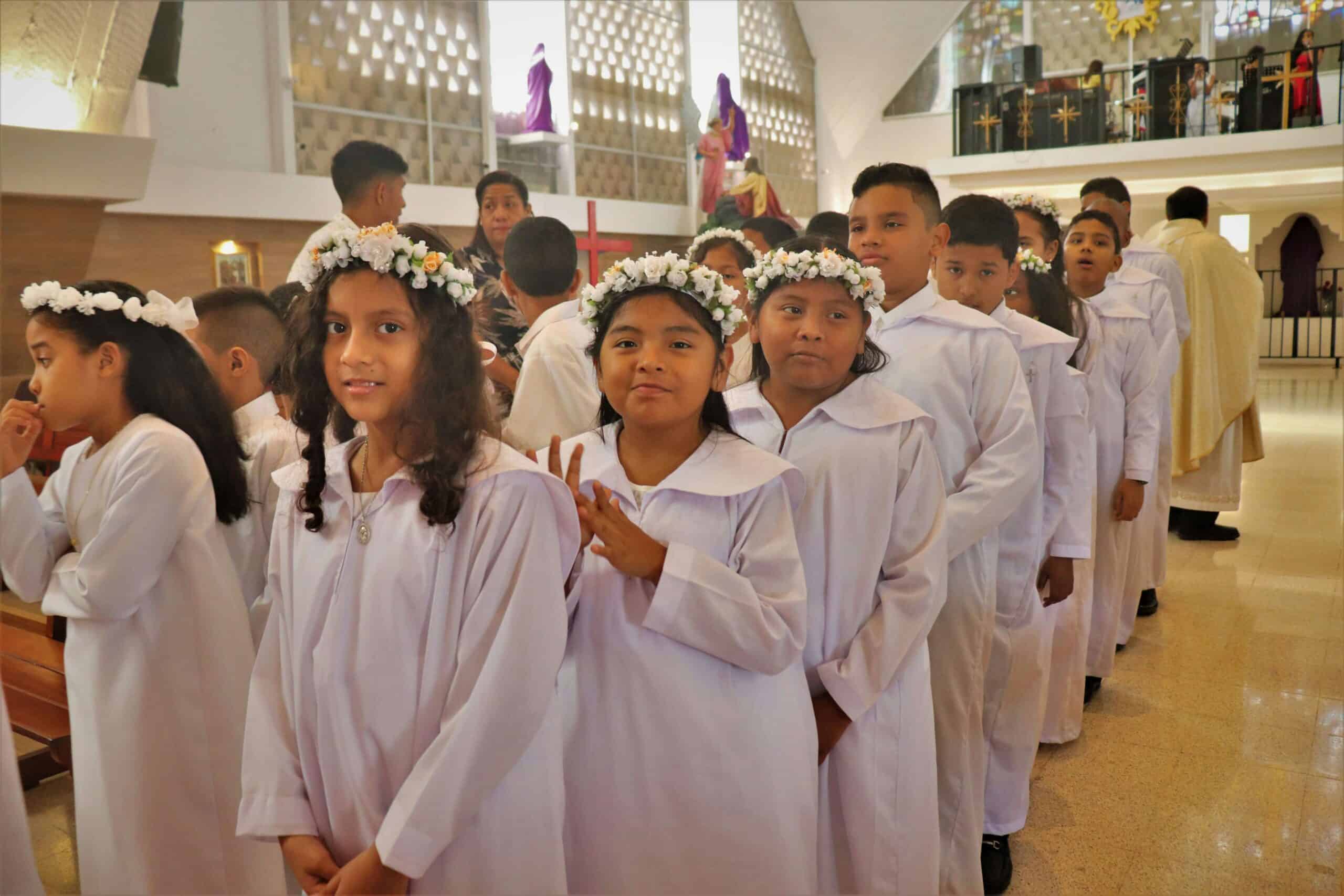 Children in white robes and flower crowns line up in a church interior, preparing for a religious ceremony. A person and statues are visible.