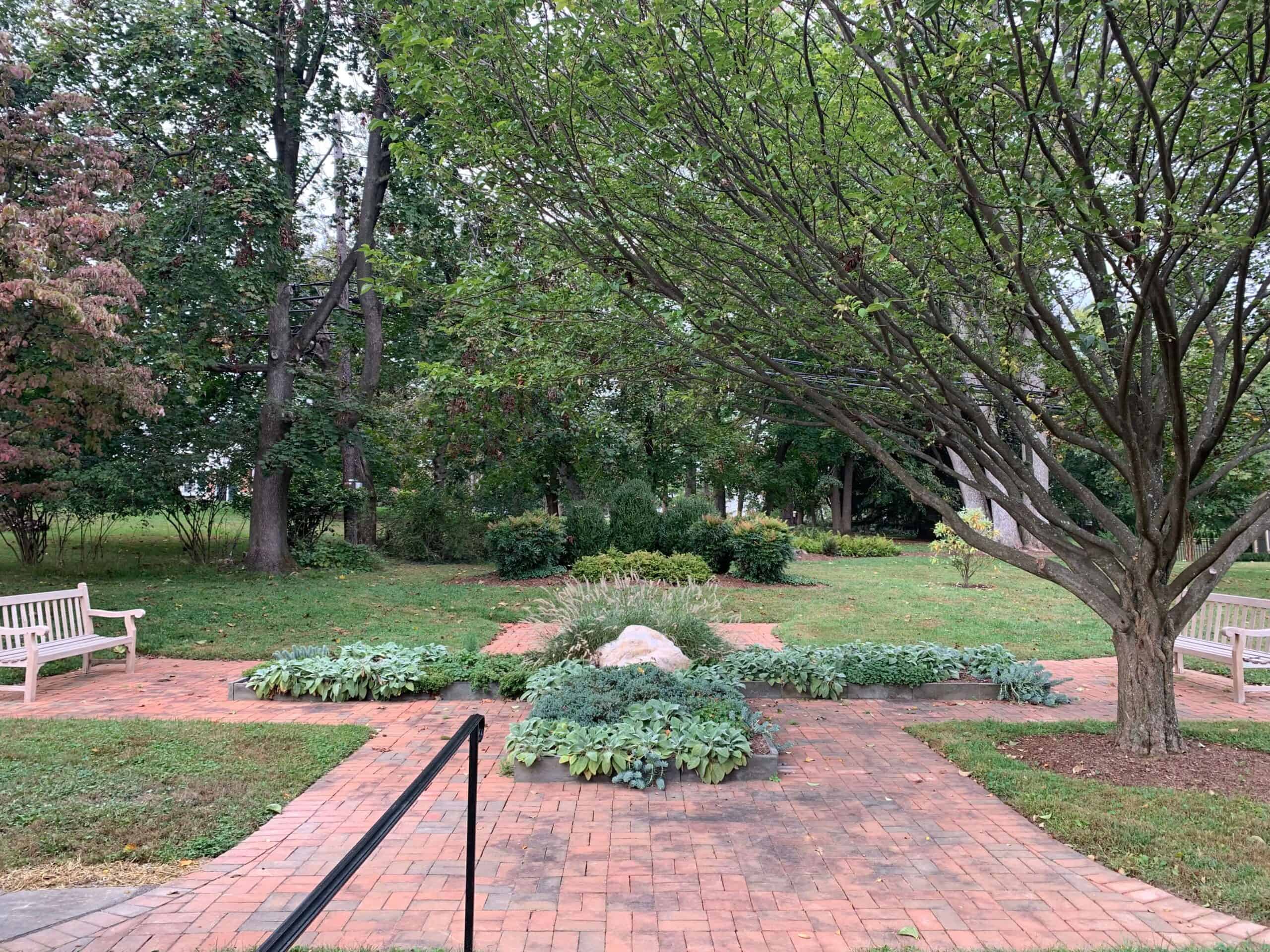 A tranquil garden with brick paths, benches, and lush greenery surrounded by trees. No landmarks or historical buildings are visible.