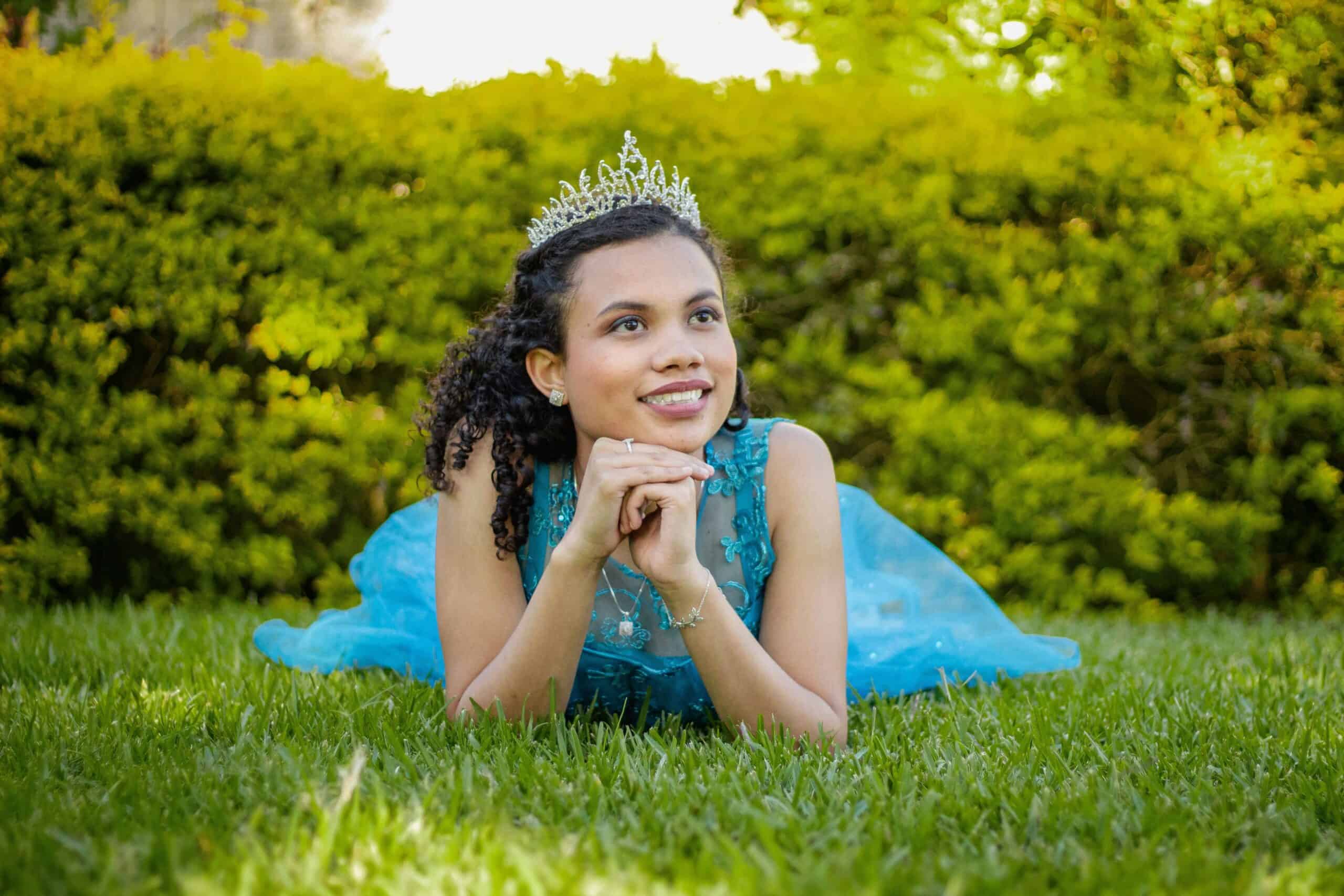 A person in a blue dress and tiara smiles while lying on grass, surrounded by vibrant greenery.