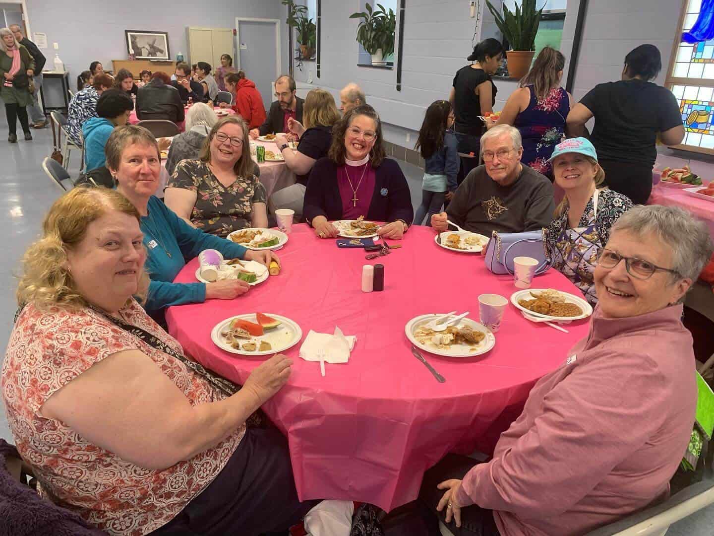 A group of people enjoys a meal at a pink-clothed table in a community gathering, with others dining in the background.