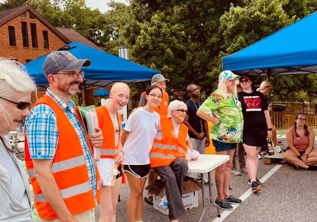 A group of people, some wearing orange vests, gather under blue canopies in front of a brick building, enjoying an outdoor event.