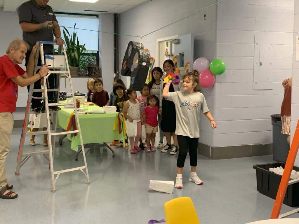 Children gather in a decorated room, excitedly watching a piñata activity. Two people assist by adjusting the setup using a ladder.