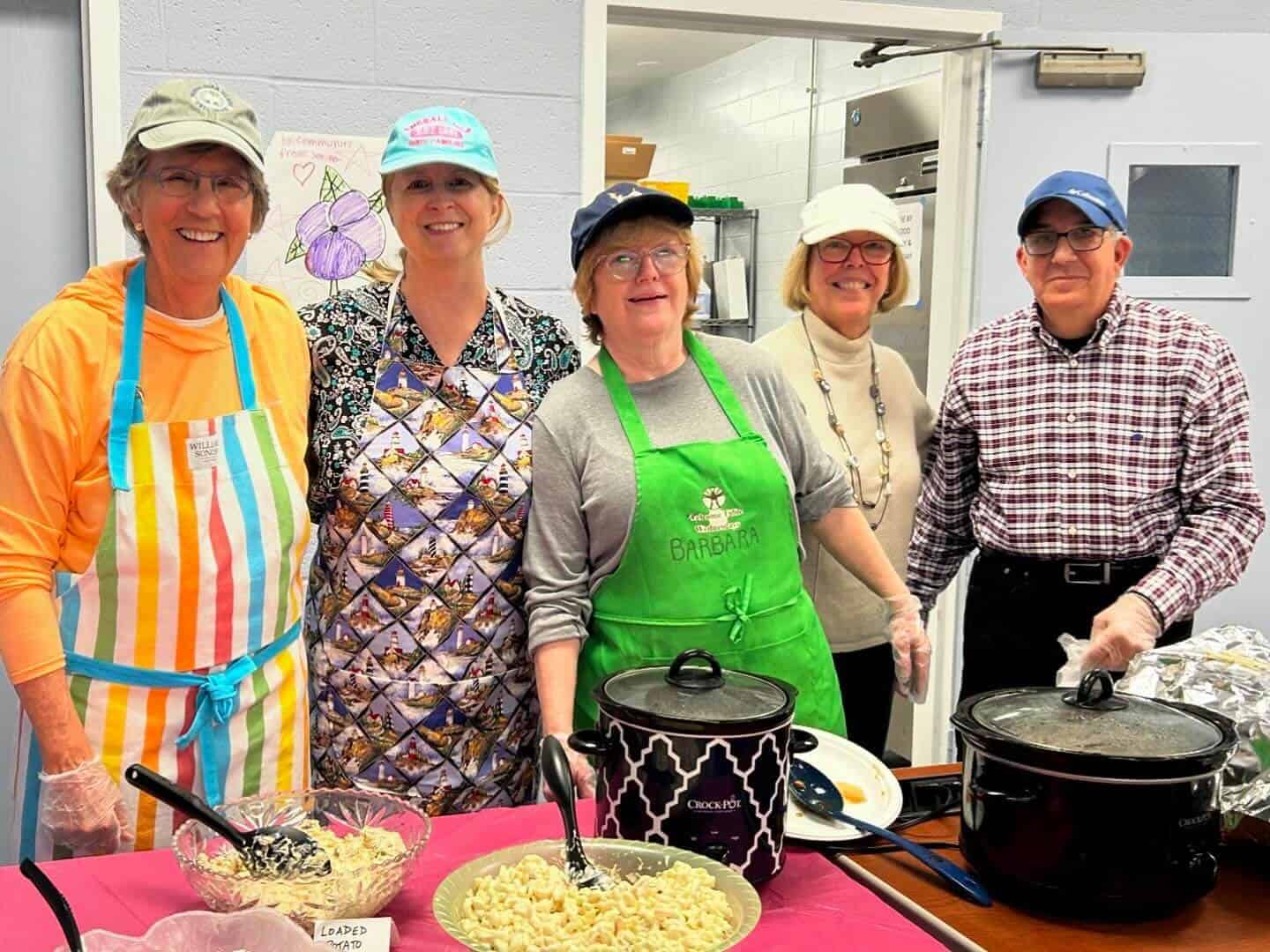 Five people smiling and wearing aprons stand behind a table with food dishes and crockpots. They're in a casual indoor setting.