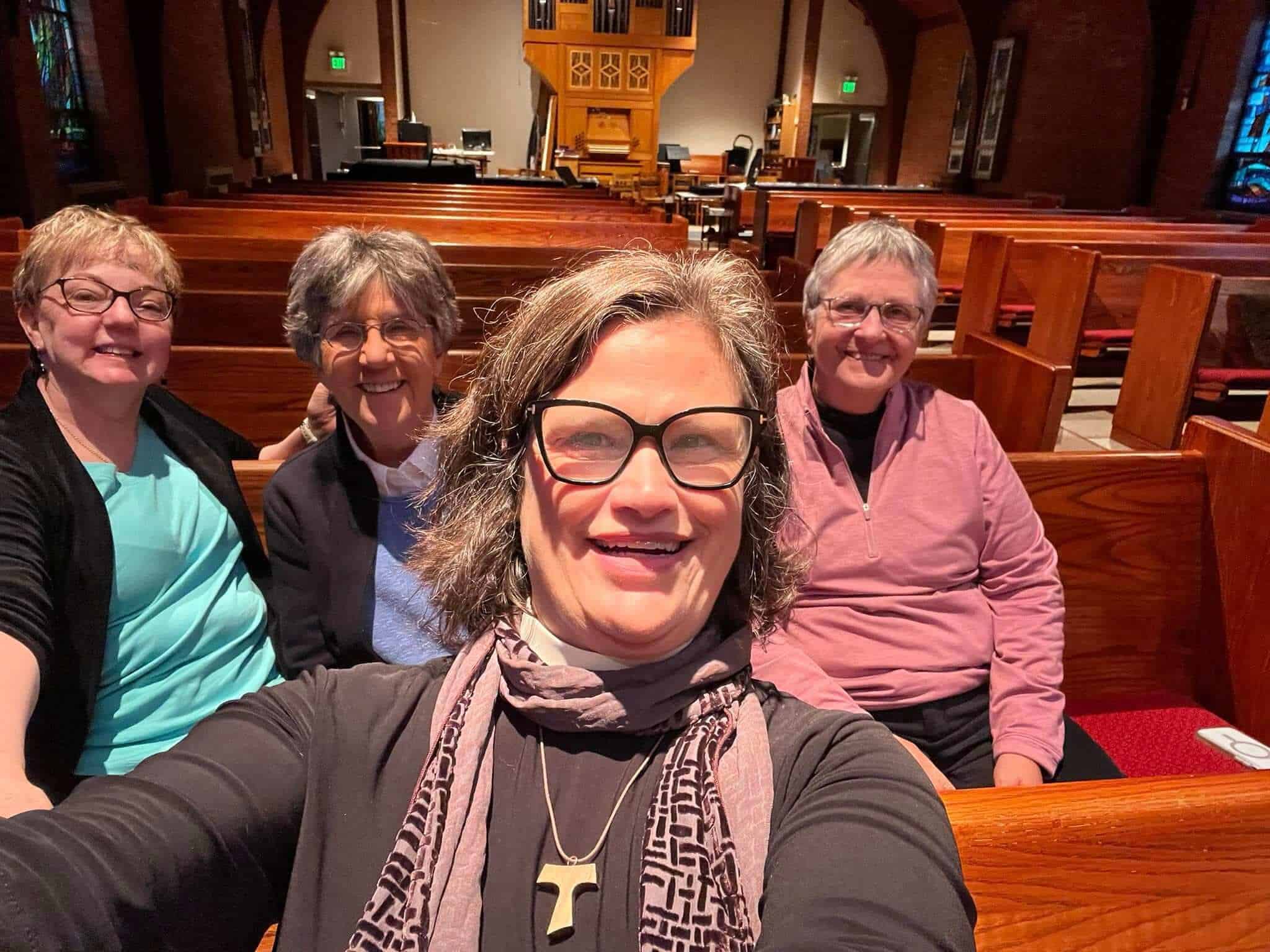 Four people sitting in wooden pews, smiling inside a church with stained glass windows and an organ in the background.