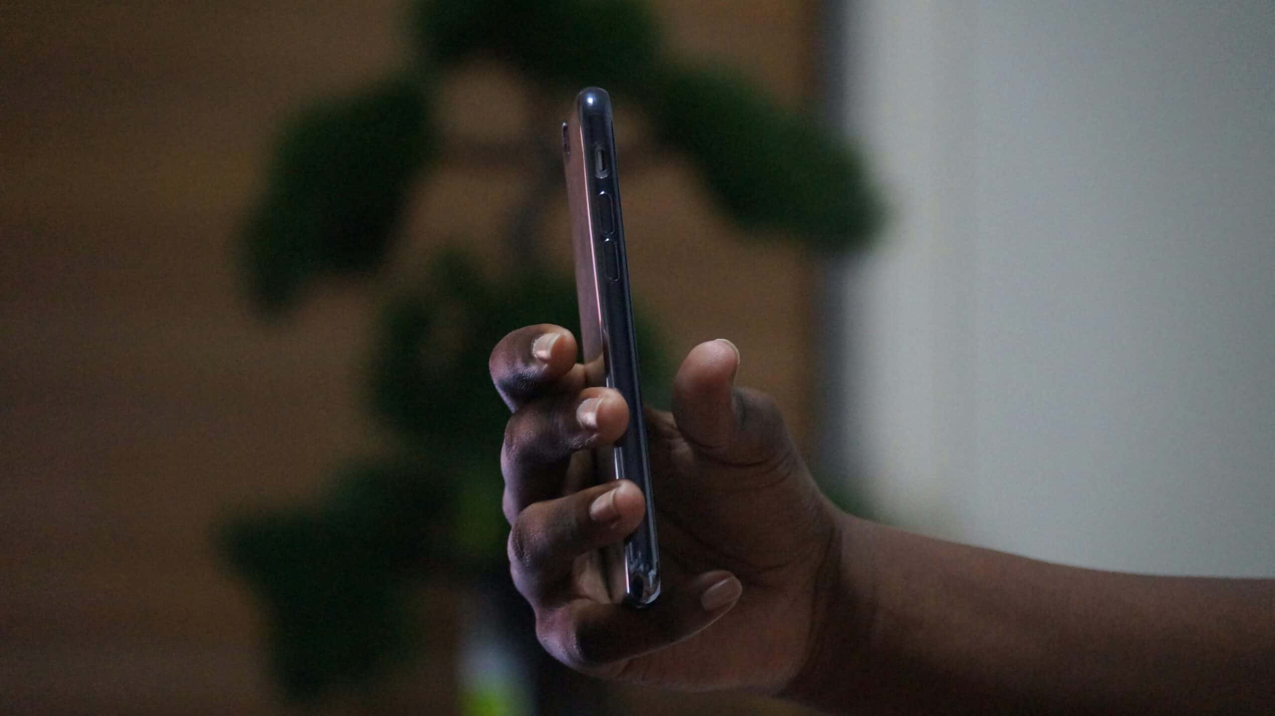 A person holds a smartphone in a relaxed grip, with a blurred background of a green potted plant and neutral wall.