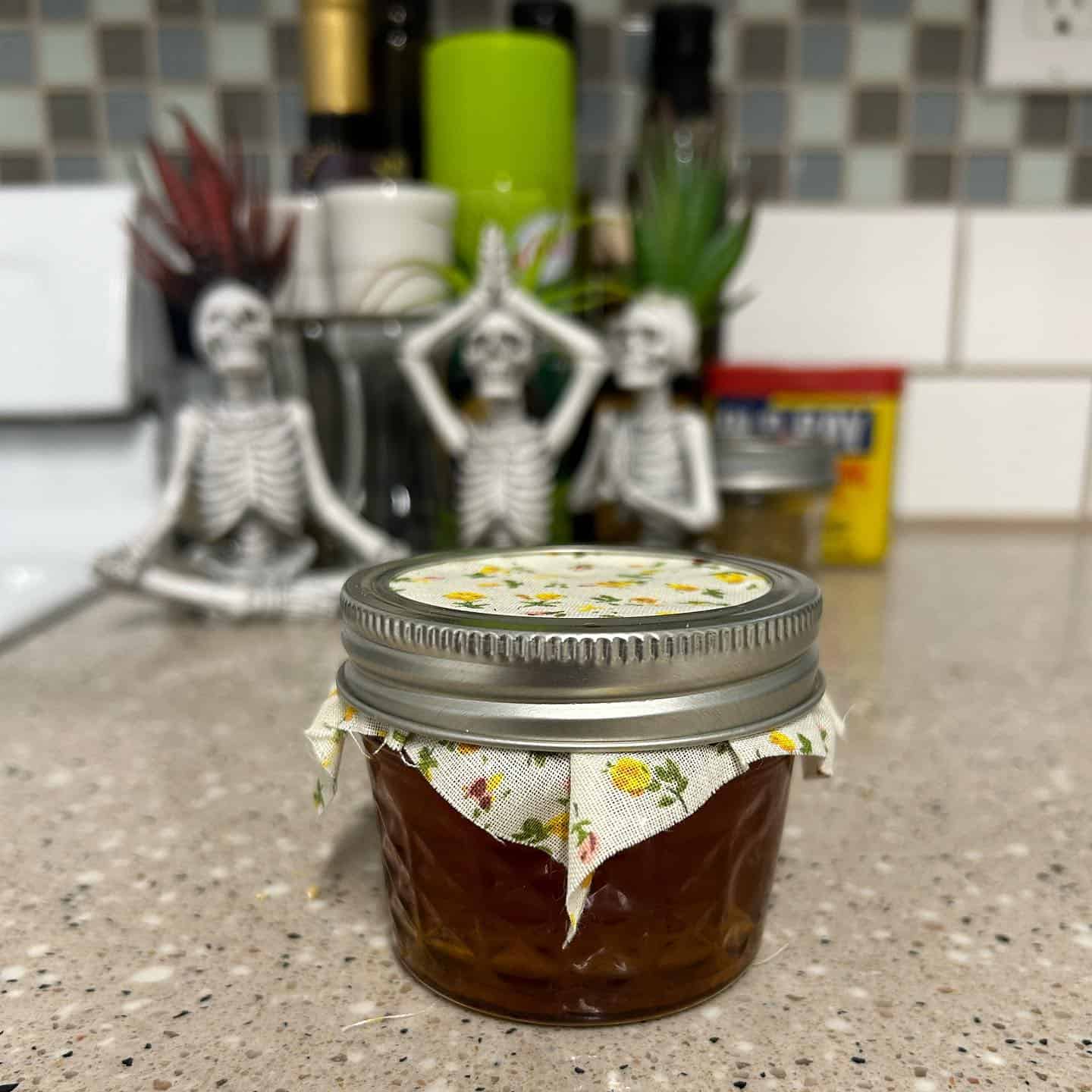 A small glass jar with floral cloth cover sits on a kitchen counter, surrounded by decorative skeleton figures in yoga poses.