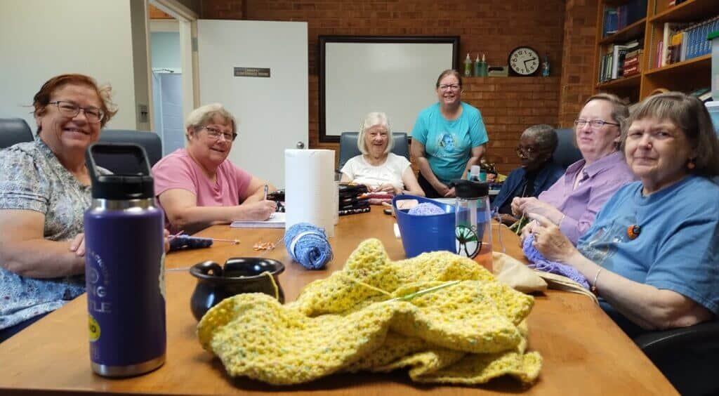 A group of people sits around a table knitting, with yarn and supplies in a cozy room with bookshelves and a clock.
