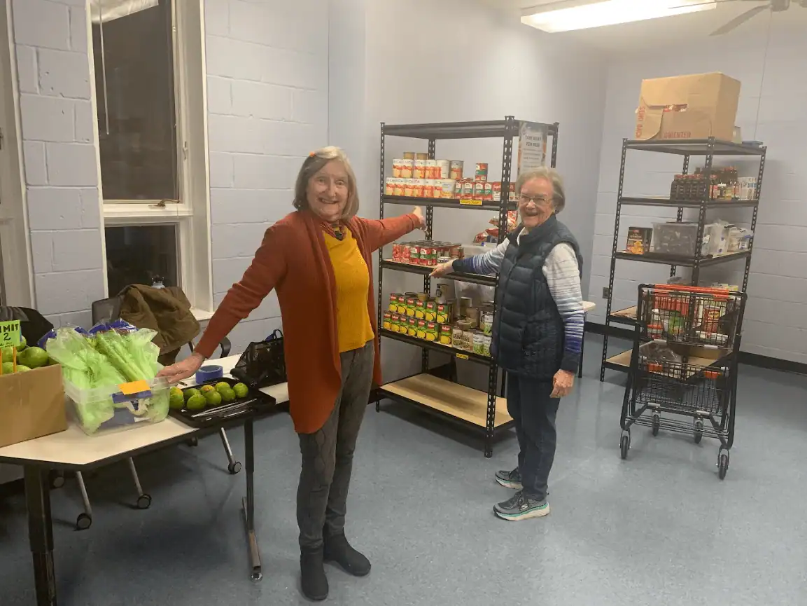 Two people smiling in a room with shelves of canned food and fresh produce on tables, suggesting a community food pantry setup.