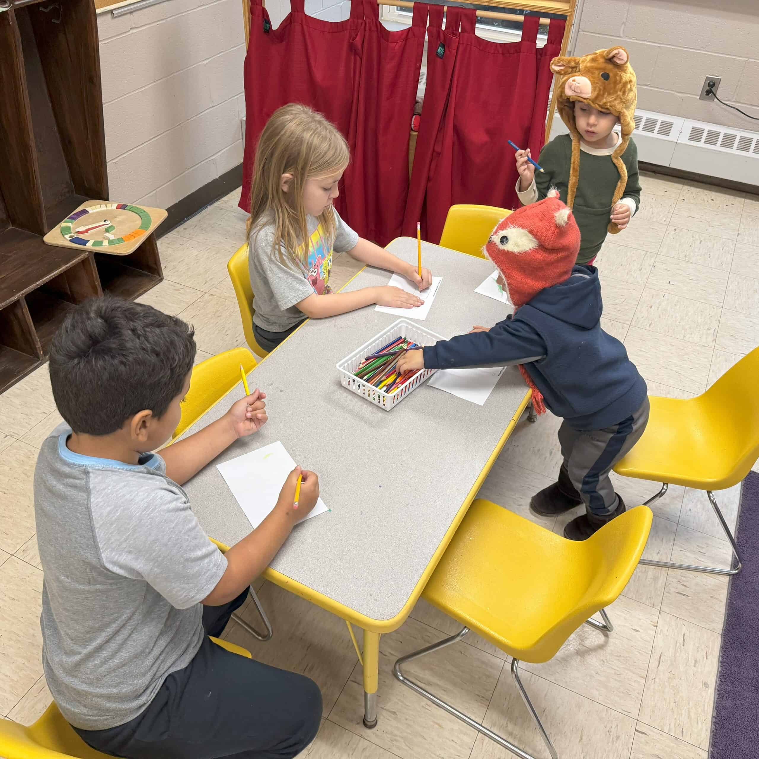 Children sit at a table in a classroom, drawing with pencils and crayons, wearing playful animal hats, surrounded by yellow chairs.
