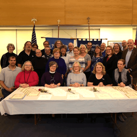 A group of people pose in a room with certificates on a table. An American flag and banners are displayed in the background.