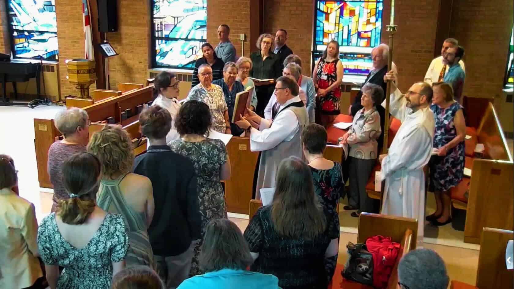 A group gathers in a church with stained glass windows. One person holds a staff, while another presents a book.